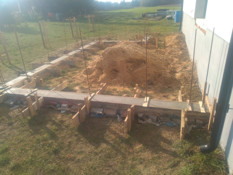 Fundamenty domu w trakcie budowy: szalunek z drewna, zalany beton, wystające pręty zbrojeniowe, pryzma ziemi w tle, widok z poziomu gruntu.