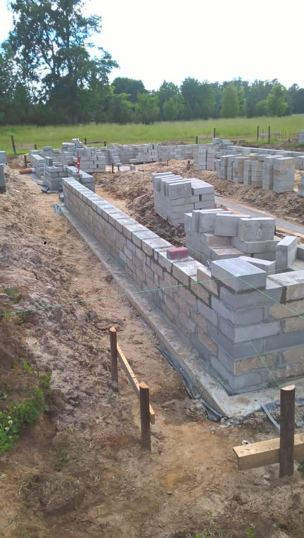 Wznoszenie muru fundamentowego z bloczków betonowych, widoczne sznurki wyznaczające poziom i pion, teren budowy w naturalnym otoczeniu z zielenią w tle.