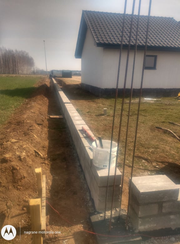 Budowa fundamentów w Koninie: Widok na częściowo wzniesioną ścianę fundamentową z bloczków betonowych, z widocznym wykopem, wystającym zbrojeniem, poziomnicą i wiadrem z zaprawą na murze.