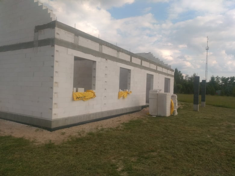 Budowa domu jednorodzinnego z białego bloczka: ściany w stanie surowym otwartym, zabezpieczone otwory okienne żółtą folią, widoczny fundament z izolacją, trawa na pierwszym planie.