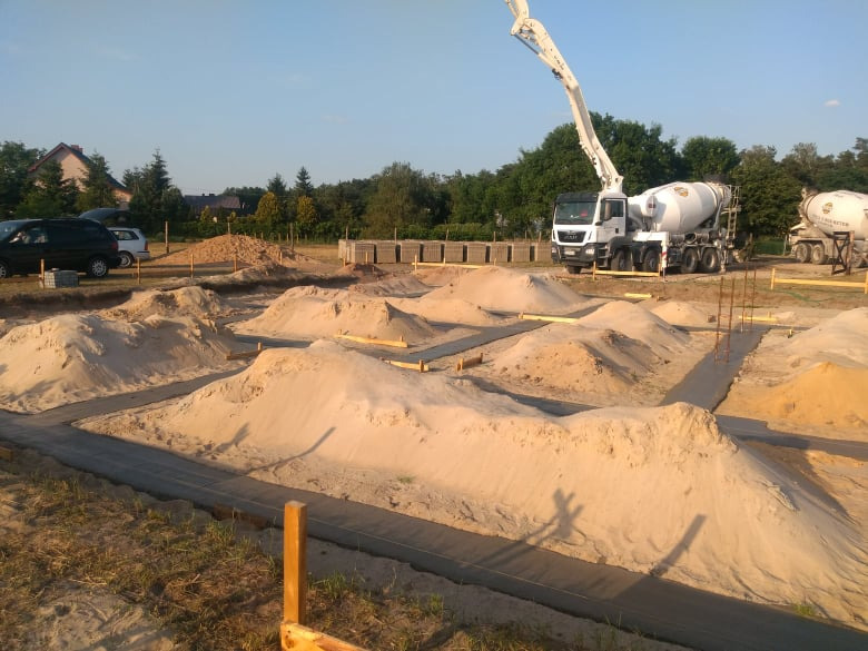 Wylewanie fundamentów pod budynek jednorodzinny, widoczny betonowy wieniec, piasek, szalunki oraz betoniarka z pompą do betonu w tle, w oddali zaparkowane samochody.