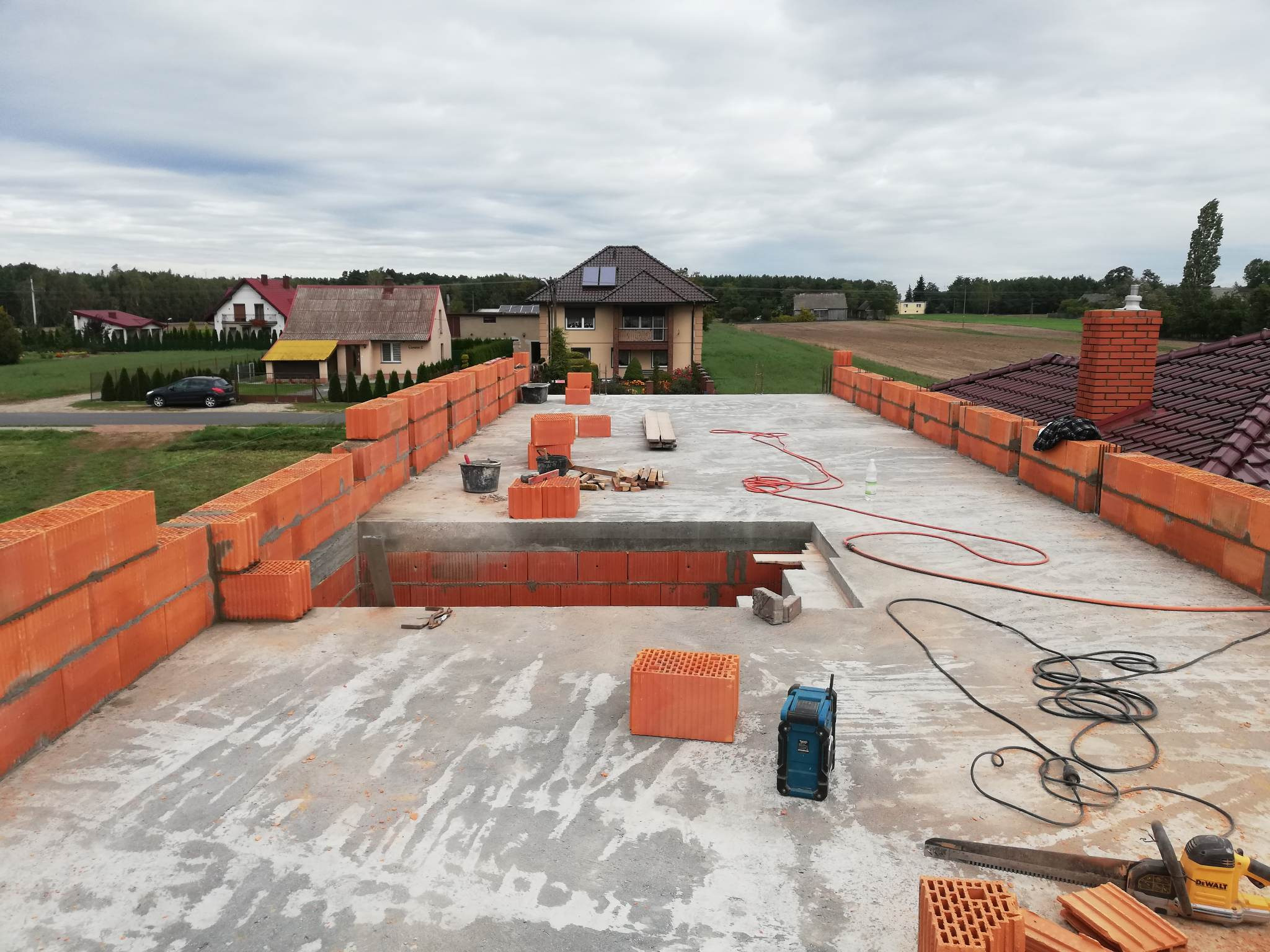 Wznoszenie muru z bloczków ceramicznych na dachu domu jednorodzinnego w trakcie budowy, widoczne narzędzia i otoczenie wiejskiej zabudowy.