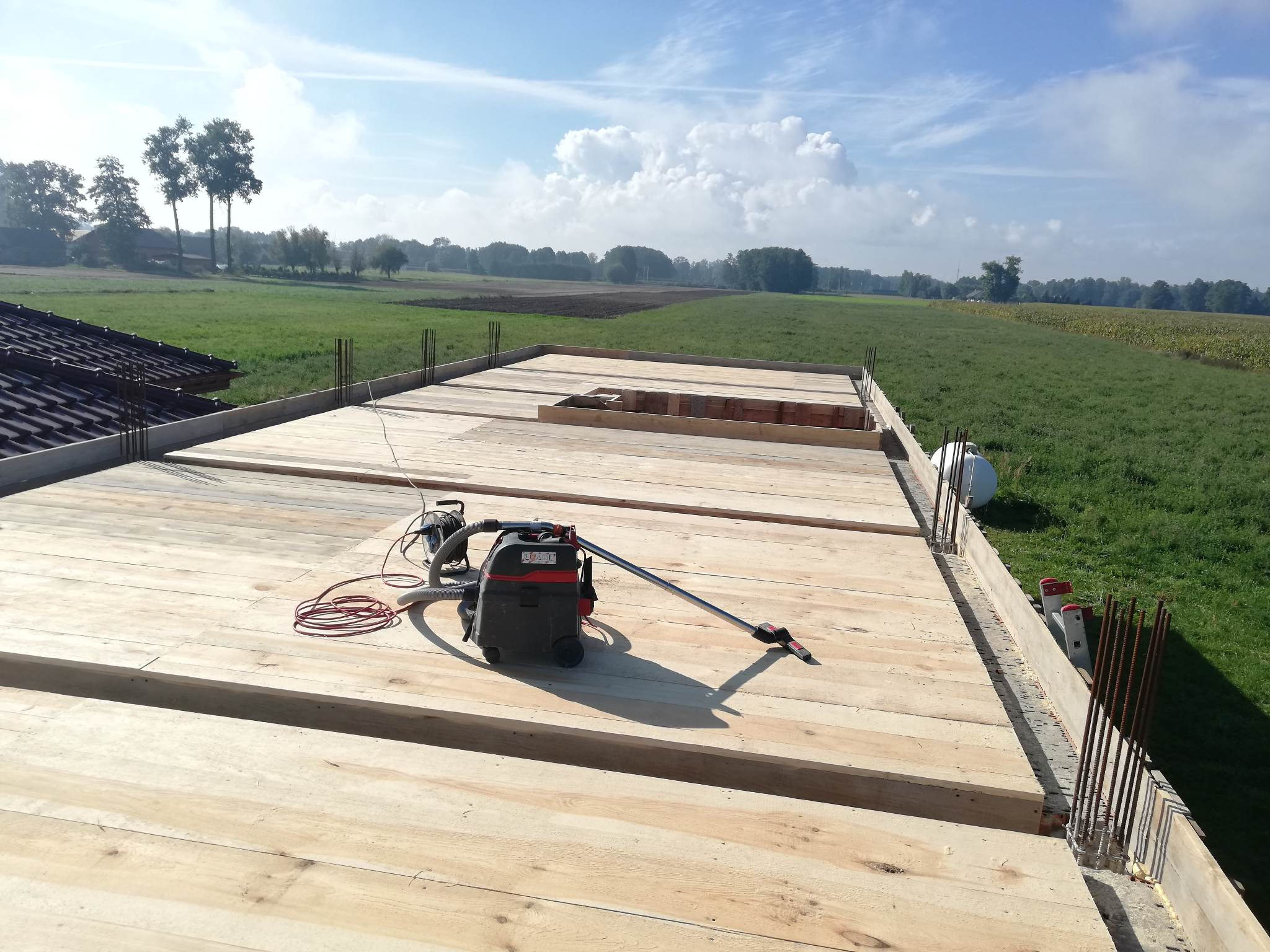 Drewniany szalunek stropu na budowie domu jednorodzinnego, widoczny odkurzacz przemysłowy, zbrojenie obwodowe i rozległe pola uprawne w tle.