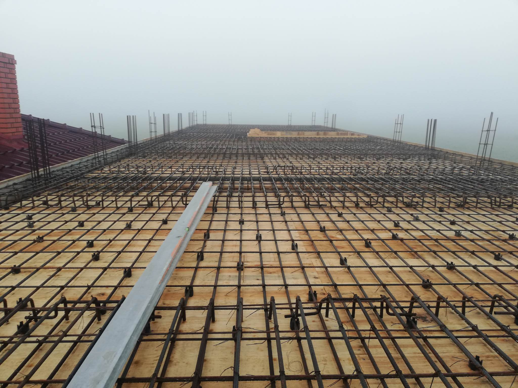 Siatka zbrojeniowa ułożona na drewnianym szalunku, widoczny poziomica, w tle ceglany komin i mgła.