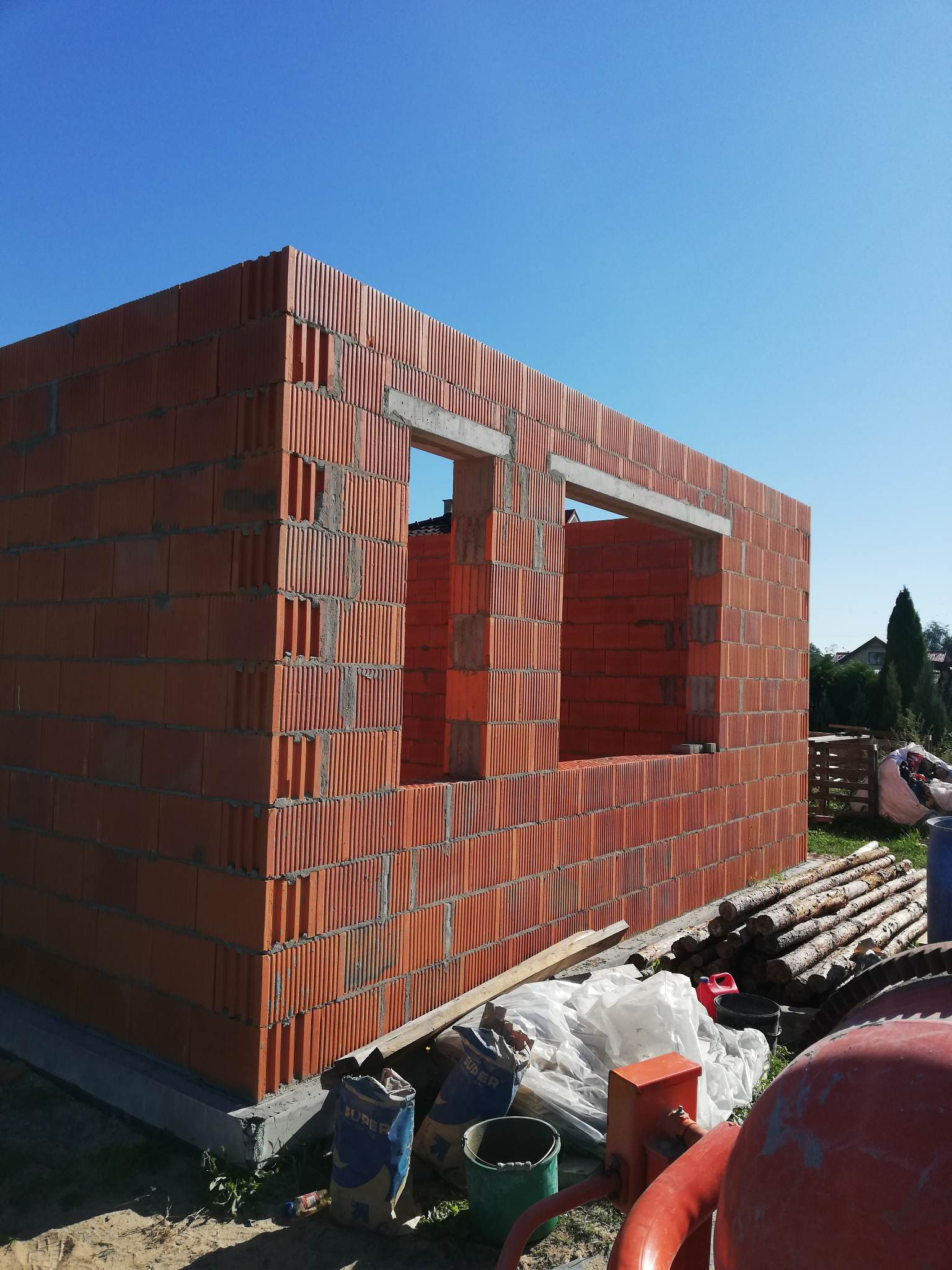 Wznoszona konstrukcja ścian domu z pustaków ceramicznych z widocznymi otworami okiennymi, na pierwszym planie worki z zaprawą i betoniarka.