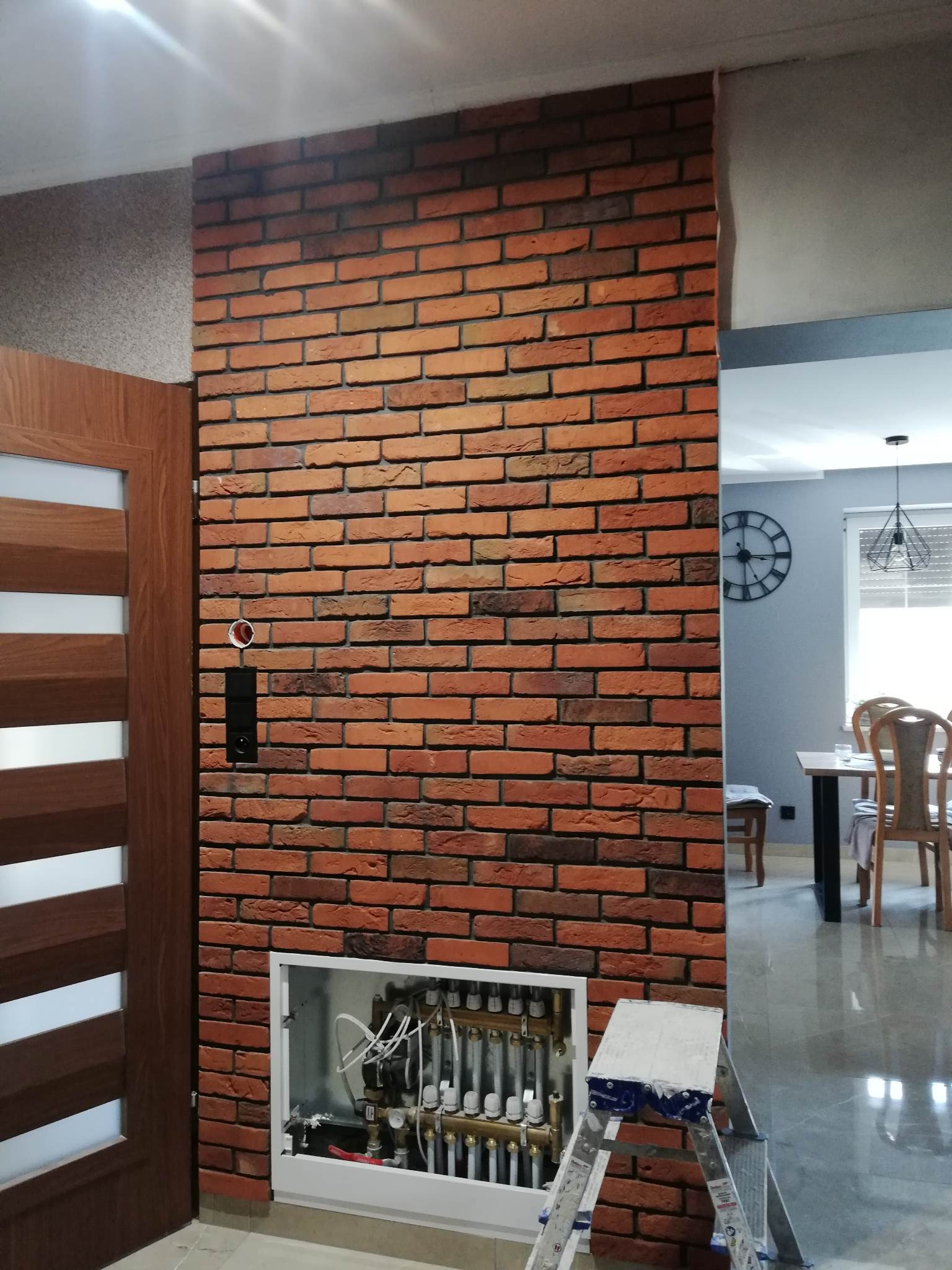 Ściana wykończona cegłą dekoracyjną w salonie, z widoczną skrzynką rozdzielacza ogrzewania podłogowego i fragmentem drabiny malarskiej. W tle widoczny salon z zegarem na ścianie.