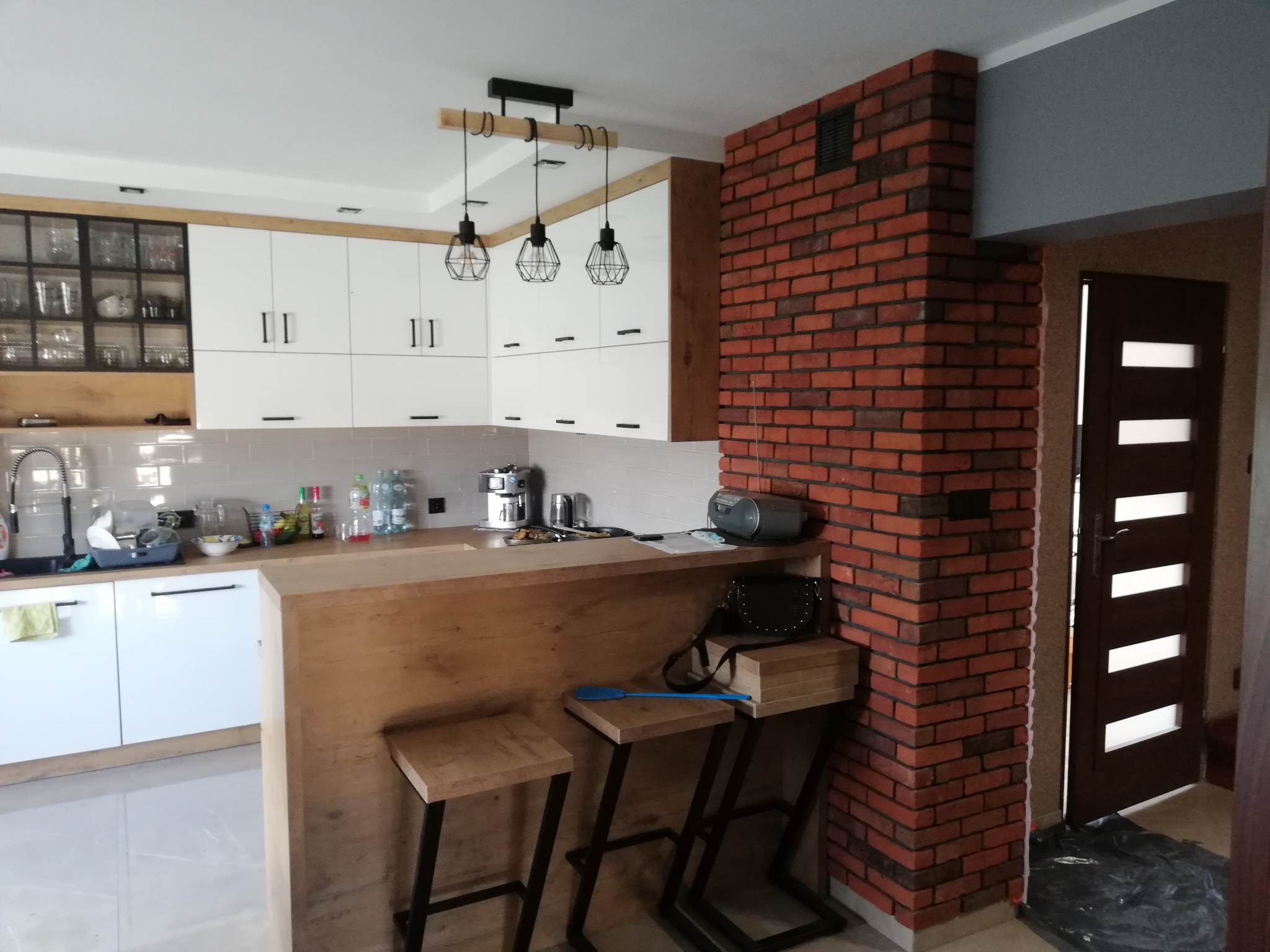 Nowoczesna kuchnia z białymi szafkami, drewnianym blatem i ceglanym filarem, oświetlona trzema wiszącymi lampami z metalowymi kloszami, widoczne dwa drewniane hokery z czarnymi nogami.
