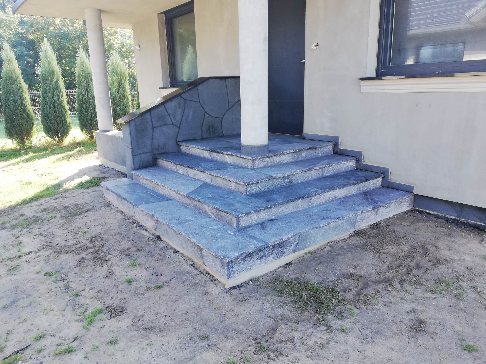 Schody wejściowe do domu wyłożone szarą kamienną płytą, z kolumną podtrzymującą zadaszenie i fragmentem elewacji w tle.