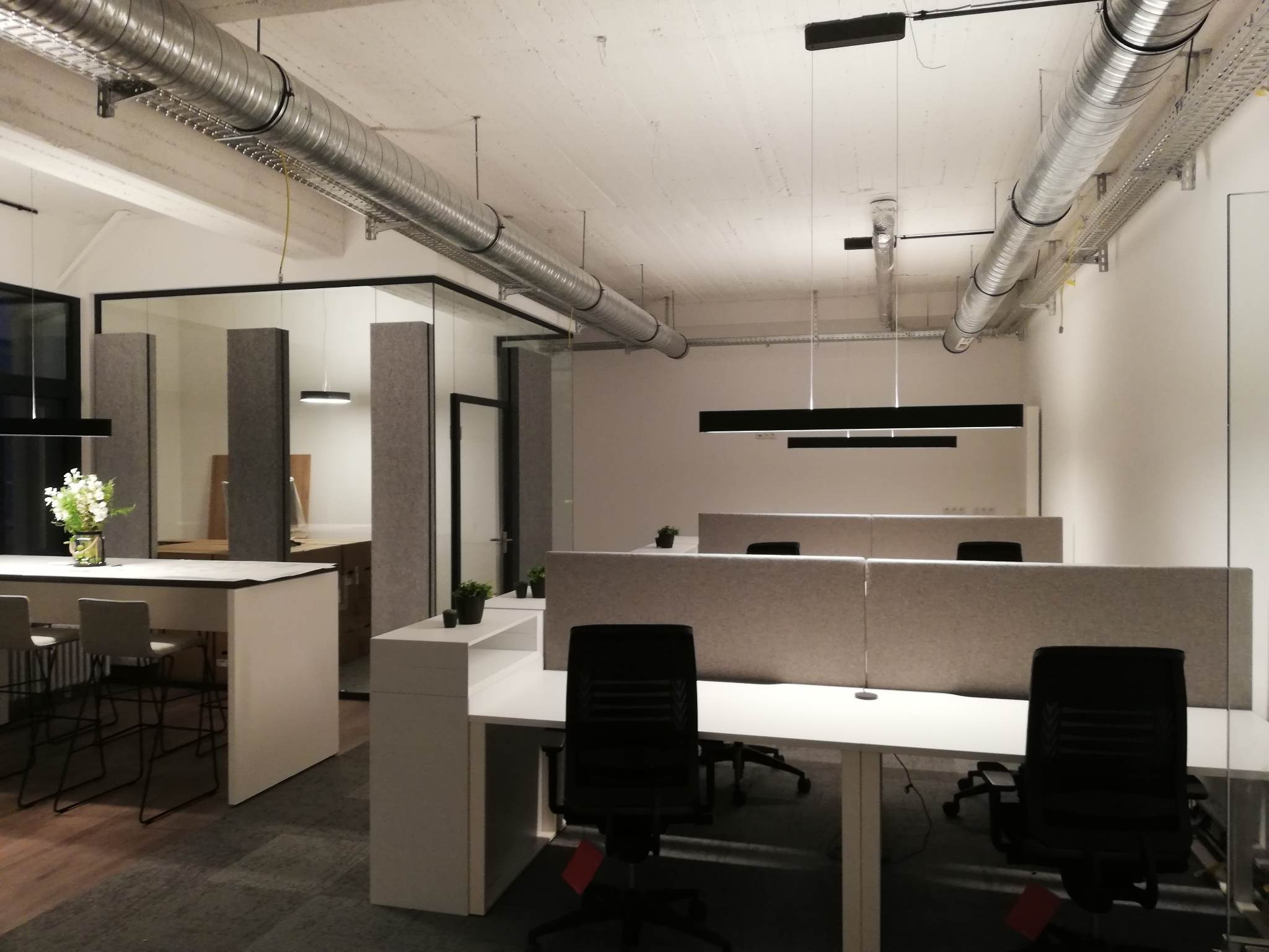 Nowoczesne biuro z białymi biurkami, czarnymi krzesłami i szklanymi ściankami działowymi, z widocznymi rurami wentylacyjnymi pod sufitem i minimalistycznym oświetleniem.