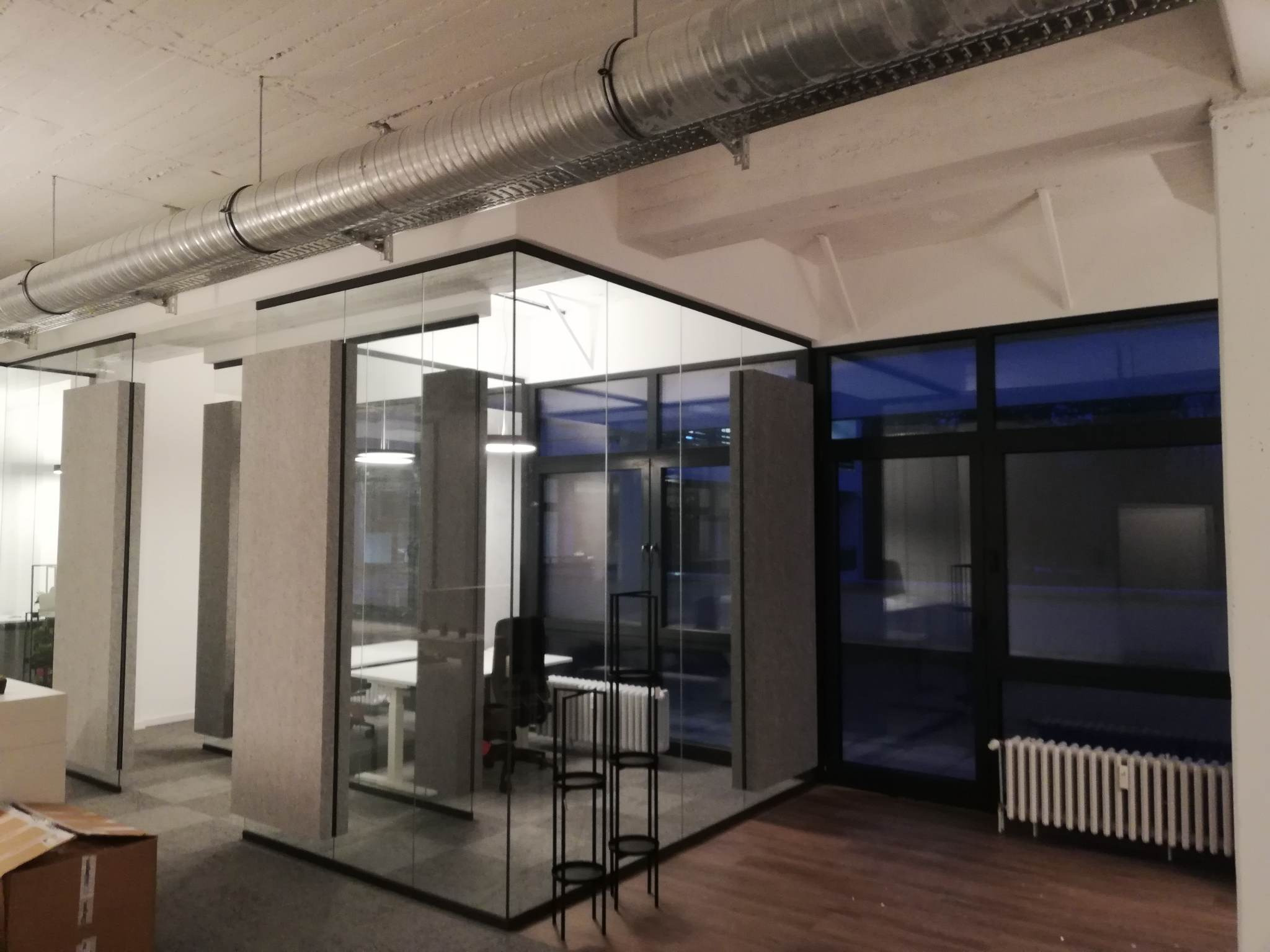 Nowoczesne biuro z przeszklonymi ścianami działowymi, widoczne biurka, krzesła i system wentylacyjny pod sufitem. Częściowo widoczne kartony sugerują prace wykończeniowe.