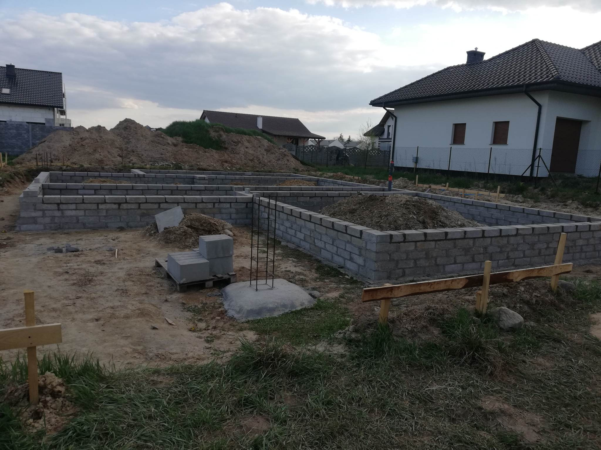 Fundamenty domu jednorodzinnego w trakcie budowy, widoczne bloczki betonowe i wystające pręty zbrojeniowe, w tle pryzma ziemi i zabudowa mieszkalna.