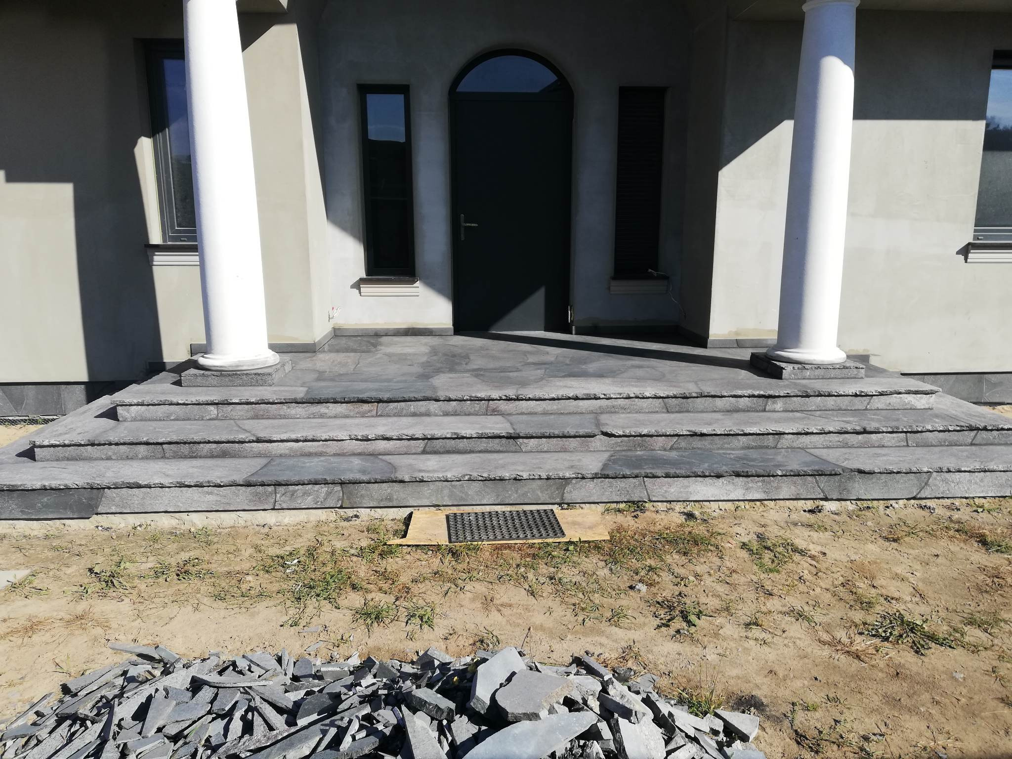 Wejście do domu z kolumnami i schodami wyłożonymi szarym kamieniem, widoczny stos kamiennych płytek przed schodami.