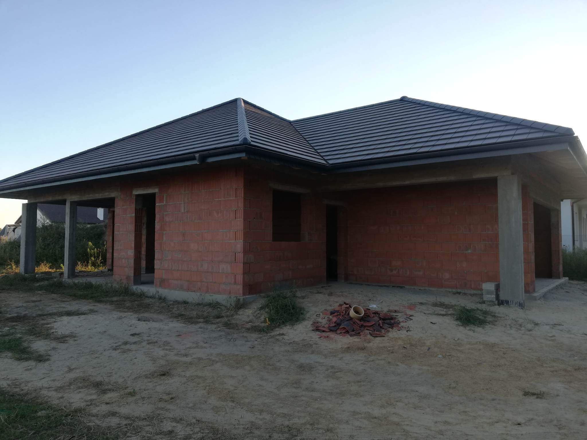 Surowy stan domu jednorodzinnego z czerwonej cegły z ciemną dachówką, widoczne niedokończone otwory okienne i drzwiowe, oraz sterta potłuczonych dachówek na piaszczystym podłożu.