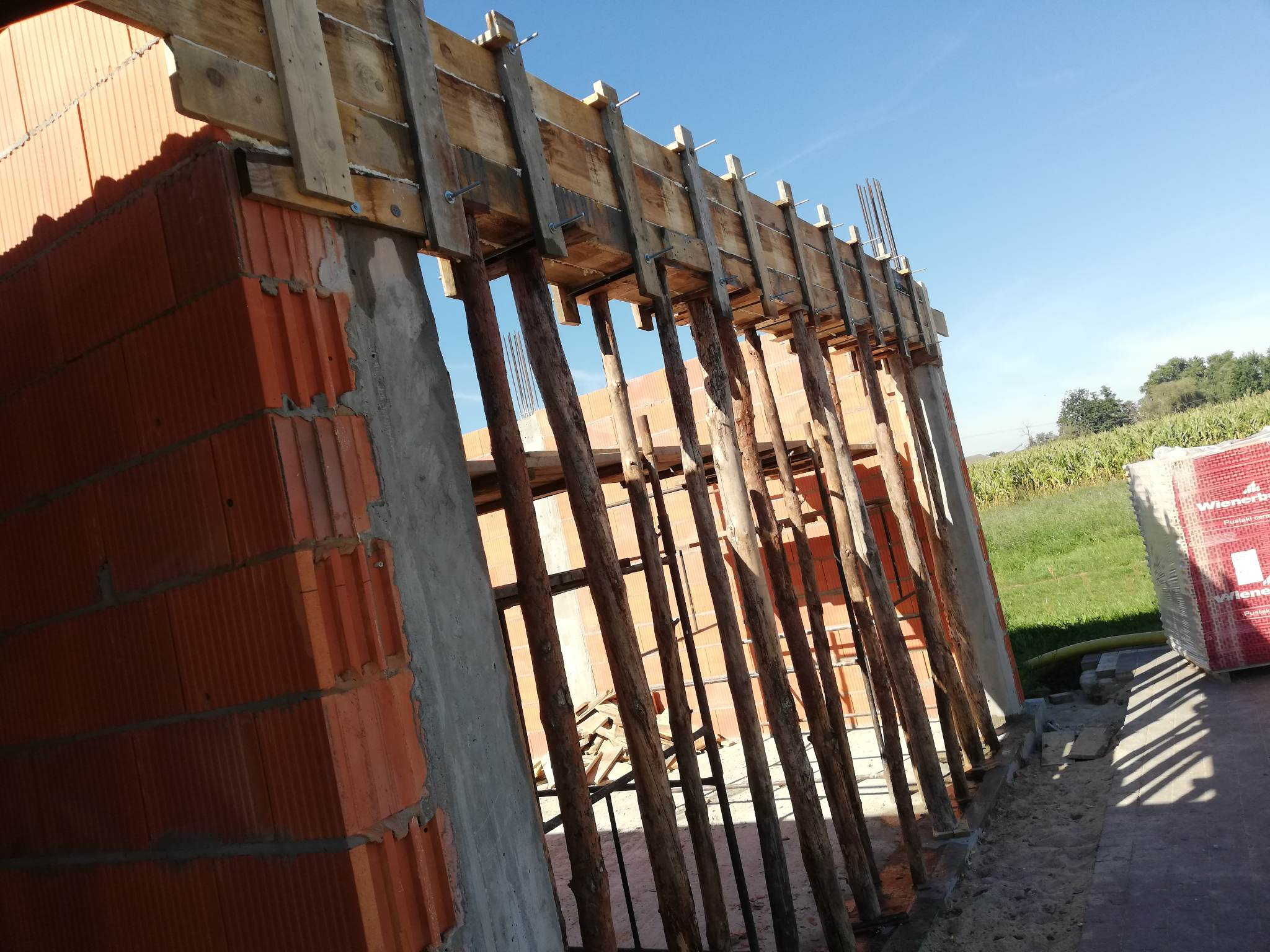 Budowa ściany z pustaków ceramicznych Wienerberger Porotherm, z drewnianym szalunkiem i podporami, widoczne zbrojenie w narożniku.