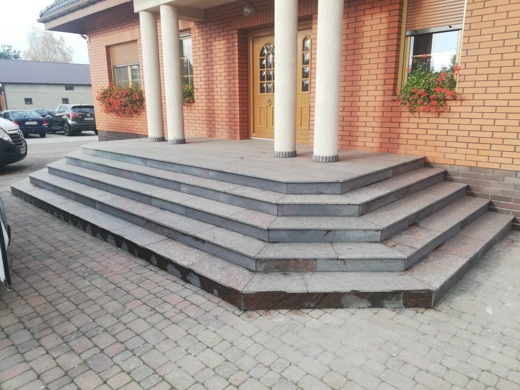 Szerokie, granitowe schody wejściowe do budynku z cegły z kolumnami, widoczne skrzynki z czerwonymi kwiatami w oknach.