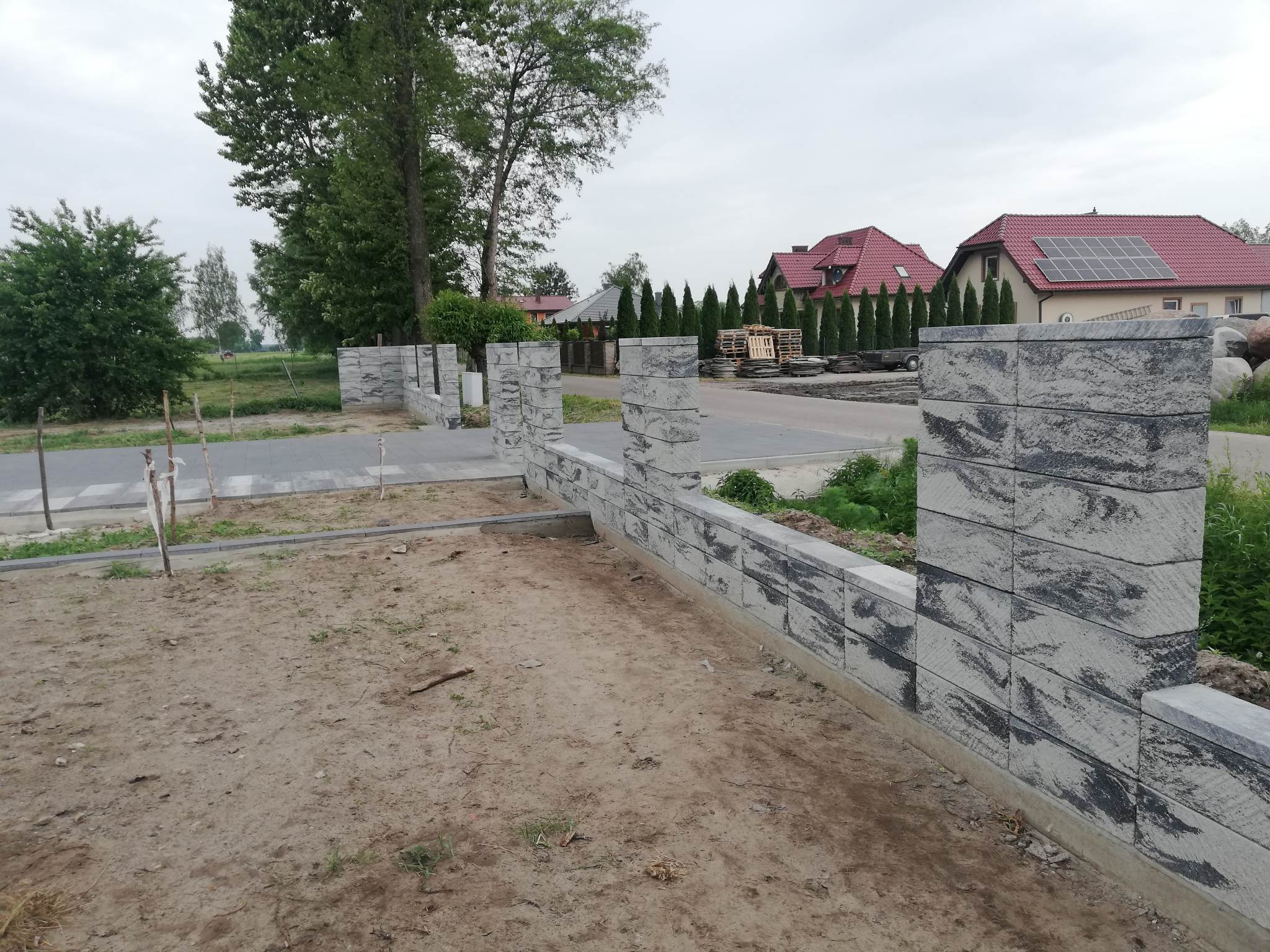 Fragment nowo budowanego ogrodzenia z szarych, betonowych bloczków o fakturze imitującej kamień, na tle domów jednorodzinnych i zieleni.