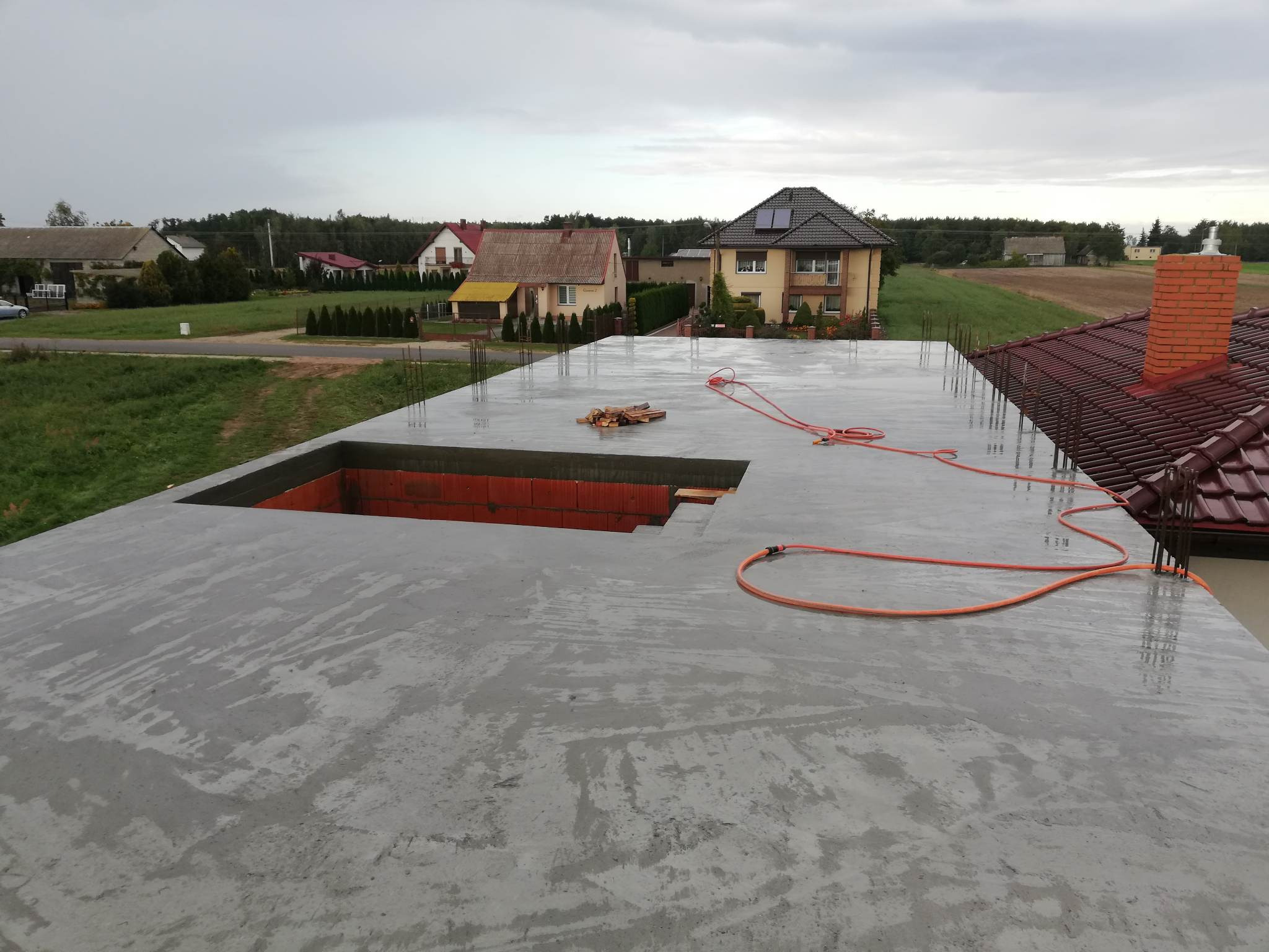 Wylany beton na stropie domu jednorodzinnego z widocznymi zbrojeniami, otworem na schody i fragmentem dachu sąsiedniego budynku w tle.