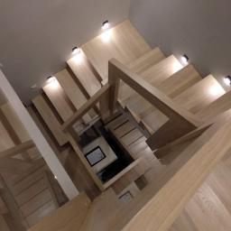 Stolarze z Warszawy - Widok z góry na drewniane schody z jasnego drewna z minimalistyczną balustradą i oświetleniem punktowym wzdłuż ściany.