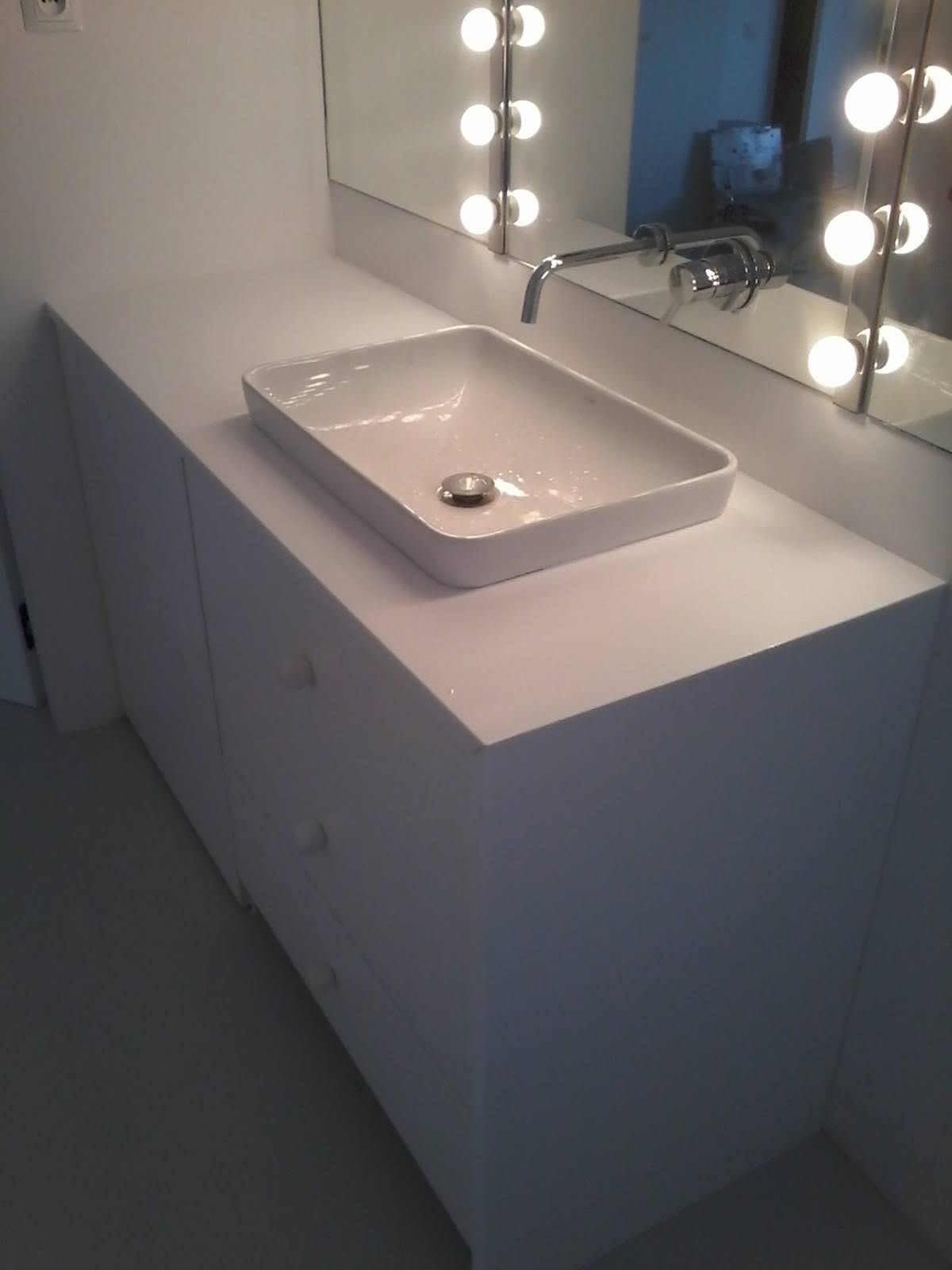 Biała, prostokątna umywalka nablatowa z chromowanym odpływem, osadzona na szafce z szufladami, oświetlona lustrem z żarówkami w stylu hollywoodzkim.