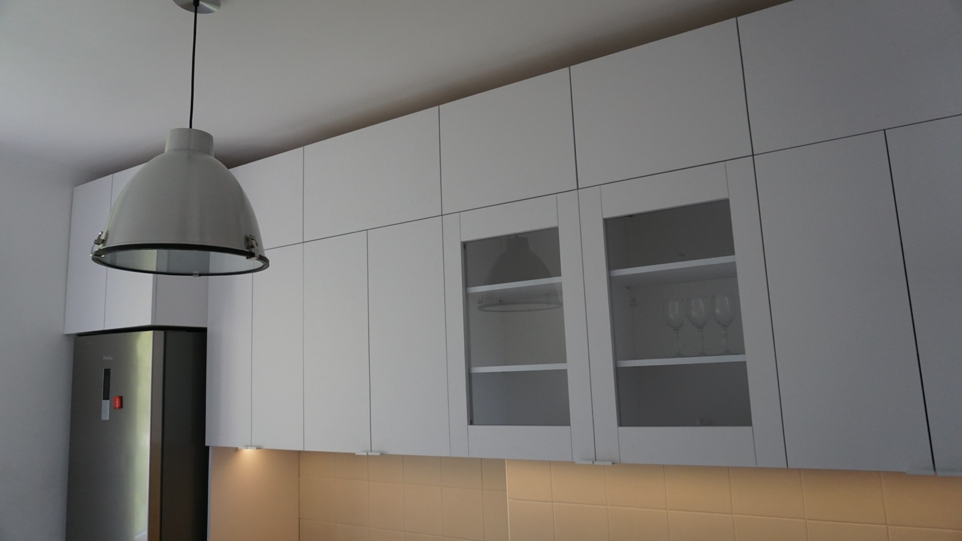 Białe, minimalistyczne szafki kuchenne podwieszane z przeszklonymi frontami, oświetlenie podszafkowe, lampa wisząca w stylu industrialnym, widoczna lodówka.
