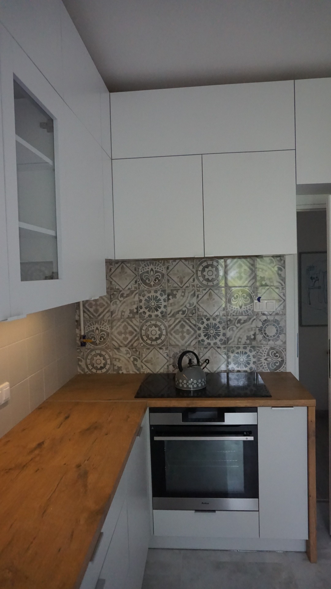 Nowoczesna kuchnia z białymi szafkami, drewnianym blatem, wzorzystymi płytkami nad indukcją i czajnikiem na niej.