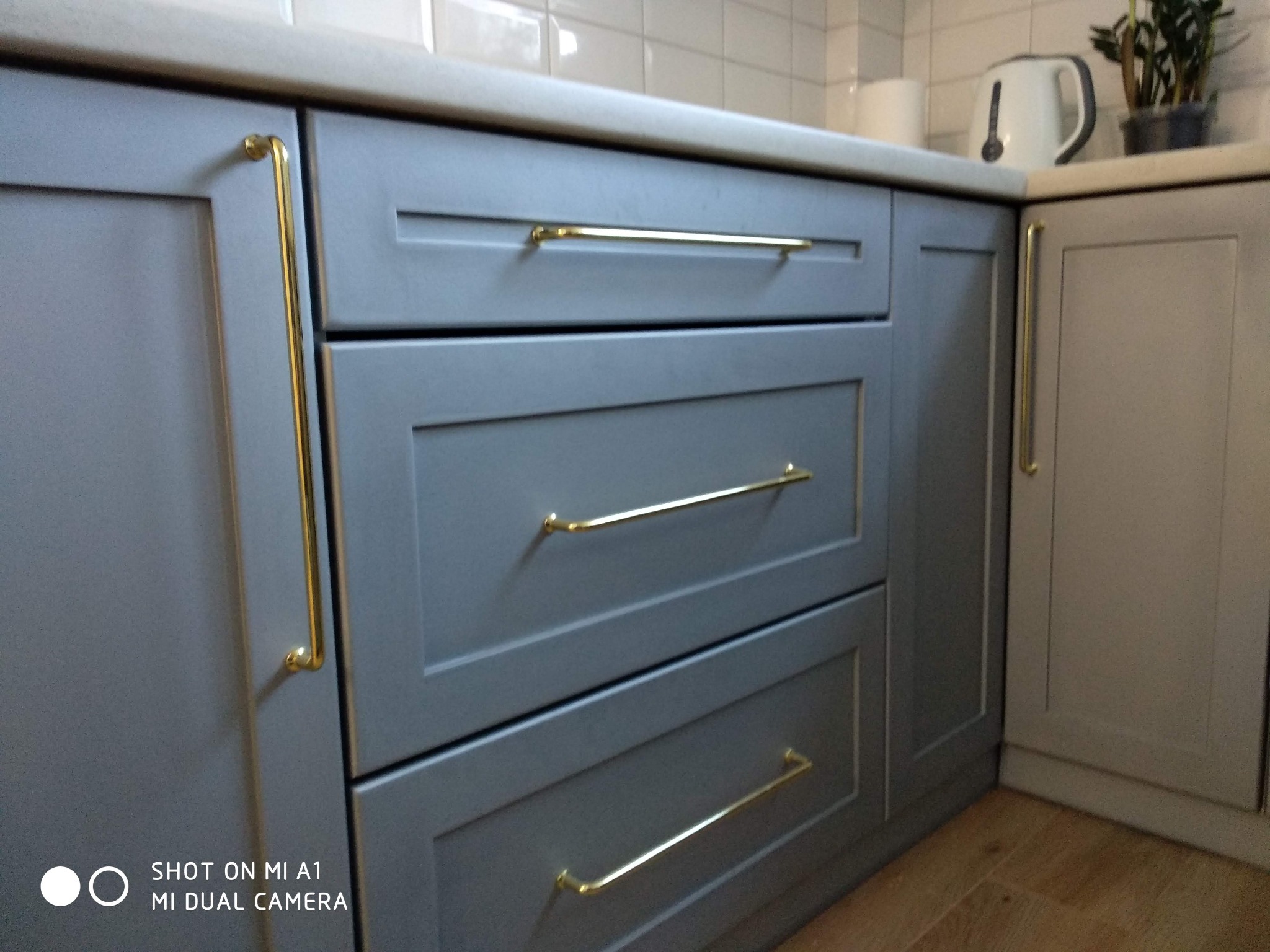 Szare szafki kuchenne z frezowanymi frontami i złotymi uchwytami, uzupełnione białymi płytkami na ścianie i akcesoriami kuchennymi w tle.