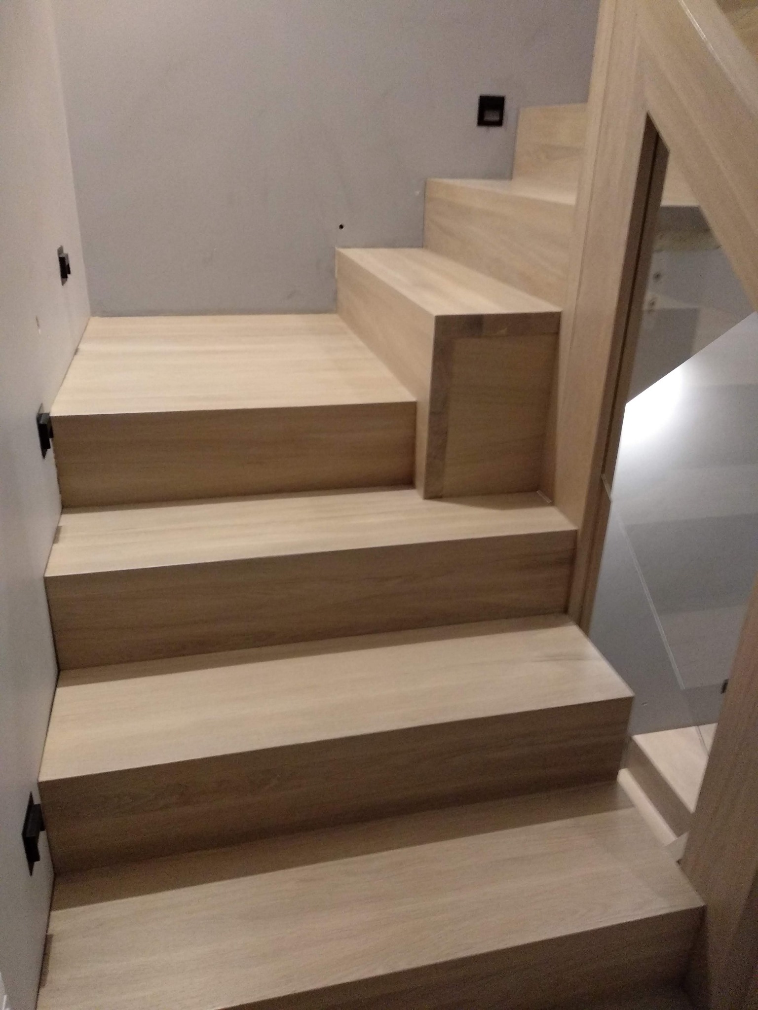 Jasne, drewniane schody z minimalistyczną balustradą ze szkła, oświetlenie punktowe na ścianie, nowoczesny design.