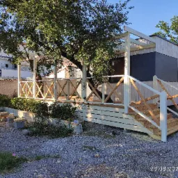 Gotowy, drewniany dom modułowy z tarasem i schodami, zlokalizowany na działce z żwirowym podłożem, w tle budynek mieszkalny.