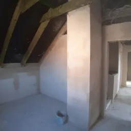Świeżo otynkowane poddasze z widocznymi drewnianymi elementami konstrukcyjnymi dachu i wiadrem po farbie w rogu pomieszczenia.