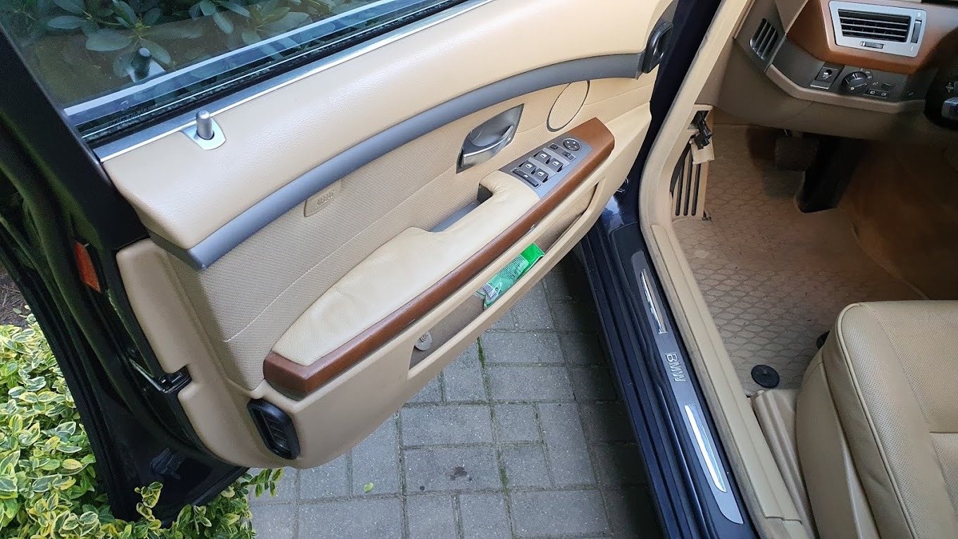 Beżowe wnętrze otwartych drzwi samochodu BMW serii 7, z widocznym panelem sterowania szybami i schowkiem na drobiazgi, na tle brukowanej nawierzchni i zieleni.