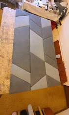 Panel tapicerowany z geometrycznym wzorem w odcieniach szarości, prawdopodobnie wykonany w Wyszkowie, leżący na stole w trakcie produkcji.