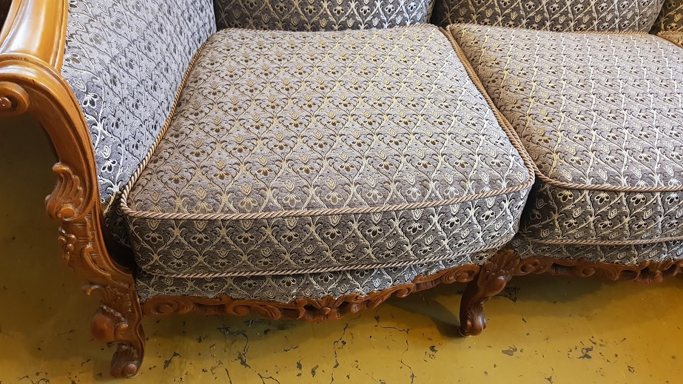 Fragment odnowionej sofy z wzorzystą tapicerką w odcieniach szarości i beżu, wykończonej ozdobnym sznurem i drewnianymi, rzeźbionymi elementami w stylu vintage.