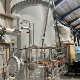 WERKKAM - Duży, szary zbiornik przemysłowy z licznymi rurami i zaworami, zabezpieczony pasami transportowymi w hali produkcyjnej. Widoczna drabina i elementy konstrukcji dachu.