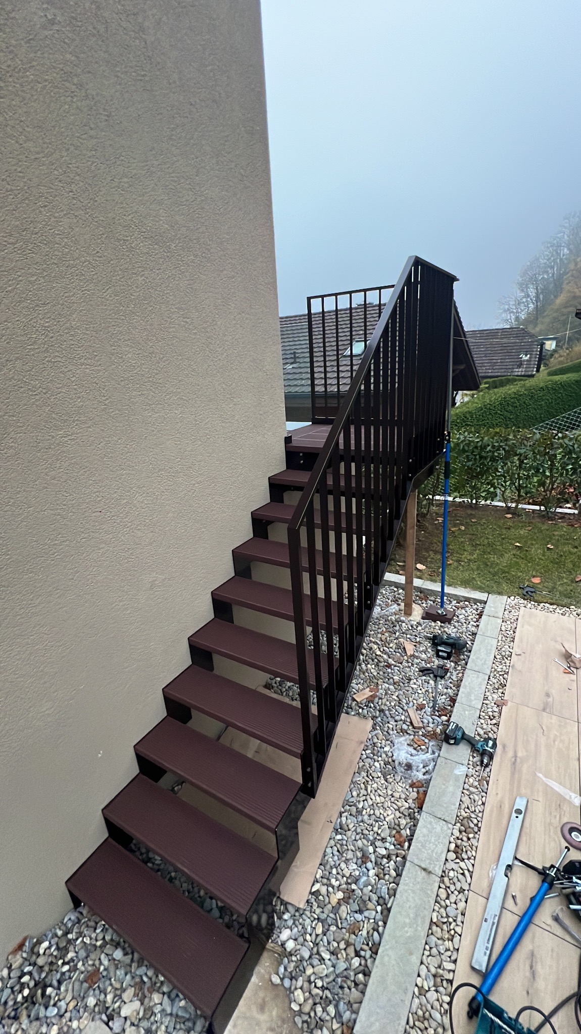 Zewnętrzne schody z brązowymi stopniami i czarną, metalową balustradą, prowadzące do ogrodu. Widoczne narzędzia i kamienie wokół schodów. Ujęcie z dołu, pochmurna pogoda.