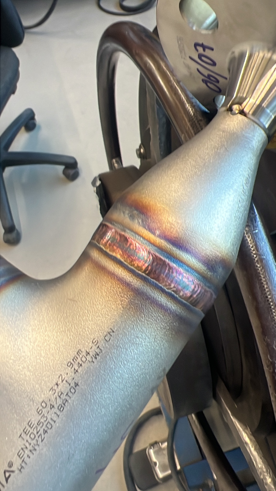 Precyzyjne spawy na metalowej rurze, widoczne tęczowe przebarwienia od ciepła, świadczące o wysokiej jakości wykonania. Detal z bliska, ukazujący profesjonalizm obróbki.