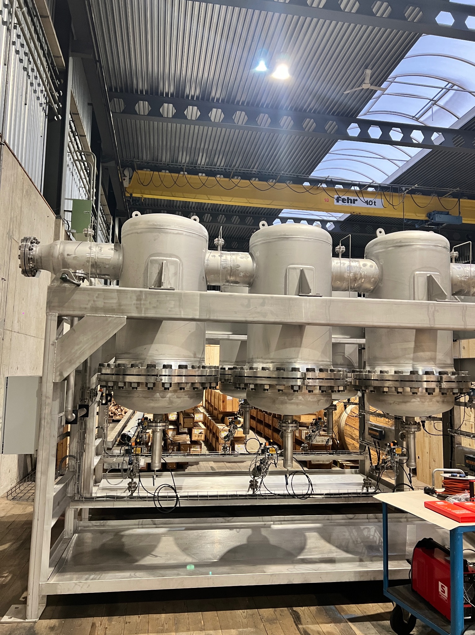 Metalowa konstrukcja z czterema zbiornikami, połączonymi rurami i zaworami, w hali przemysłowej z oknami dachowymi i suwnicą. Widoczne narzędzia na wózku.