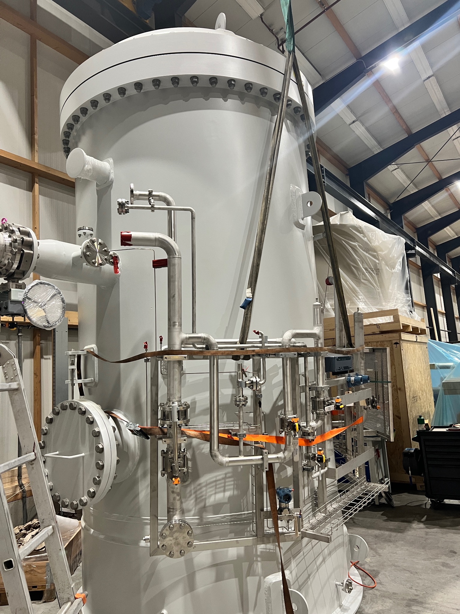 Duży, szary zbiornik przemysłowy z licznymi rurami i zaworami, zabezpieczony pasami transportowymi w hali produkcyjnej. Widoczna drabina i elementy konstrukcji dachu.