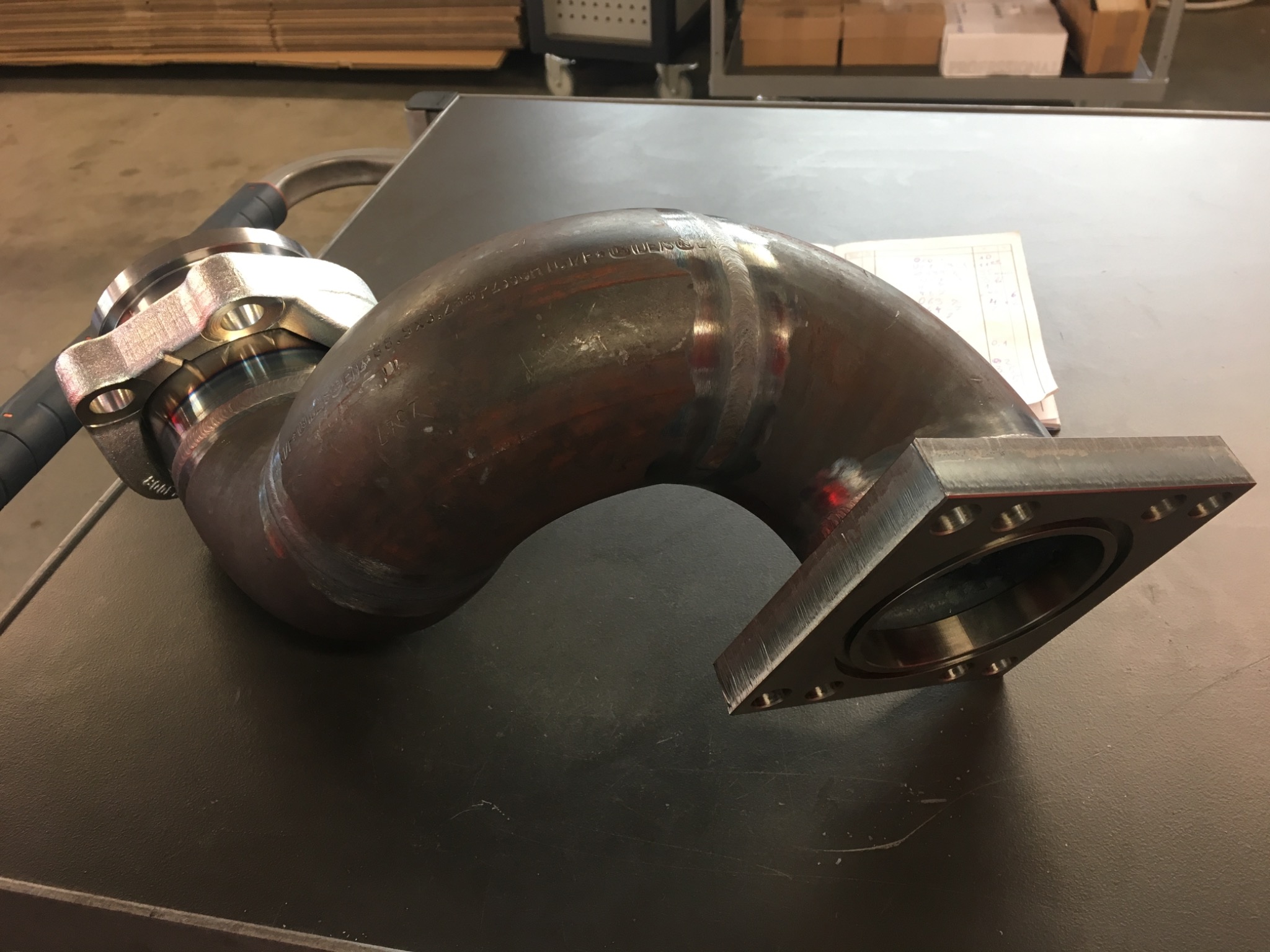 Ugięty, grubościenny element rurowy ze stali z przyspawanymi kołnierzami, leżący na stole warsztatowym. Widoczne ślady po spawaniu.