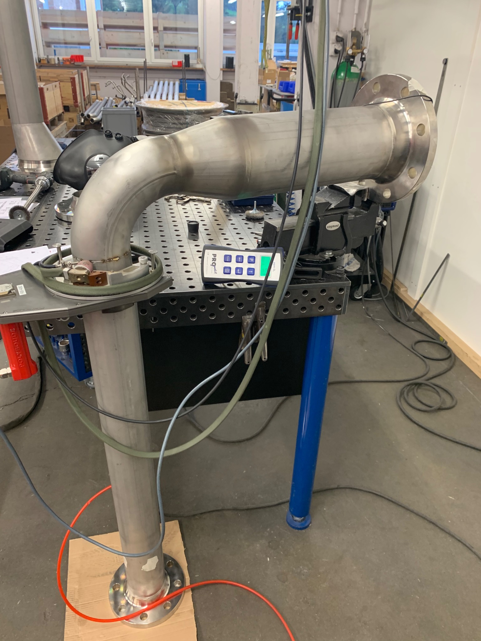 Metalowa rura z kołnierzem, spawana, zamocowana na stole warsztatowym z oprzyrządowaniem pomiarowym, widoczne spawy i ślady obróbki.