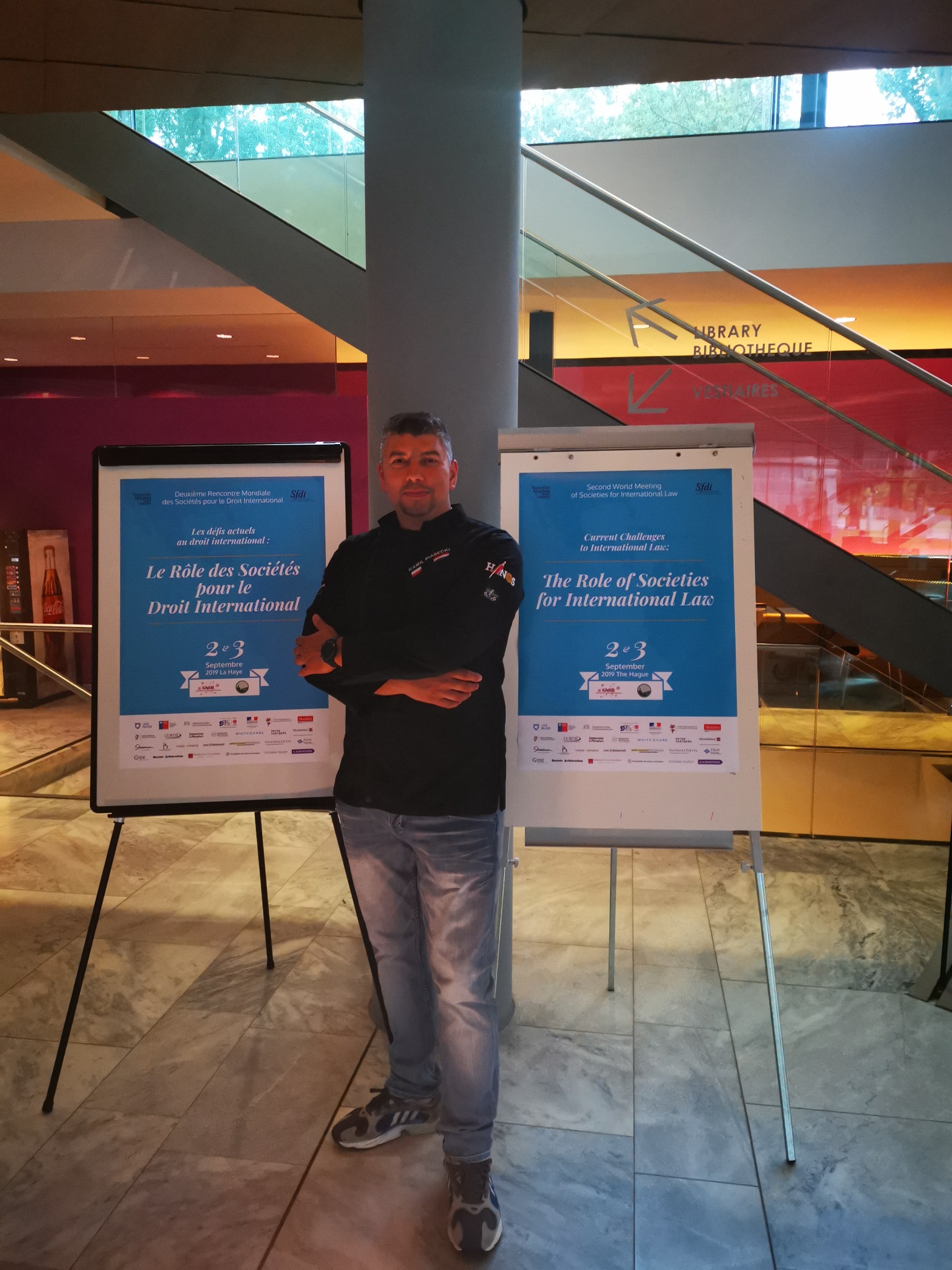 Mężczyzna w czarnej bluzie stoi na tle dwóch flipchartów z plakatami konferencji prawniczej w budynku o nowoczesnej architekturze.