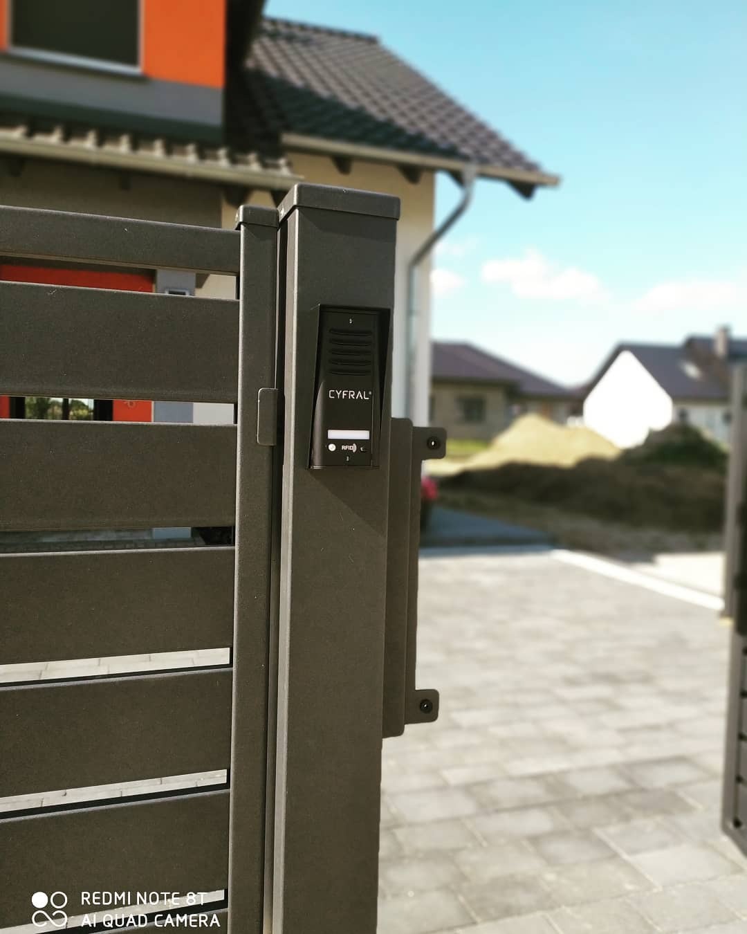 Szary panel wejściowy z czytnikiem RFID marki Cyfral zamontowany na metalowej bramie posesji. W tle widoczny dom z pomarańczową elewacją.