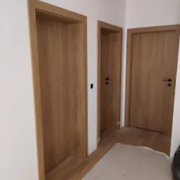 Trzy drewniane drzwi wewnętrzne z czarnymi klamkami i ościeżnicami, widoczne w korytarzu z białymi ścianami i częściowo zabezpieczoną podłogą.