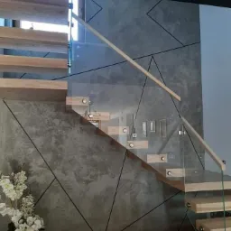 Nowoczesne schody drewniane z przezroczystą balustradą szklaną, zamocowaną na metalowych uchwytach, na tle ściany z geometrycznym wzorem w odcieniach szarości, z dekoracyjnym bukietem białych...