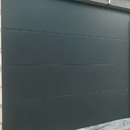 Ciemnoszara segmentowa brama garażowa w trakcie montażu, widoczne surowe mury z cegły i betonu oraz folia ochronna na dole.