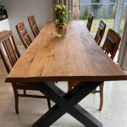 Długi, rustykalny stół z litego drewna z czarną, krzyżową podstawą, otoczony krzesłami z drewnianymi ramami i skórzanymi oparciami, z wazonem tulipanów na blacie, widok na ogród przez okno.