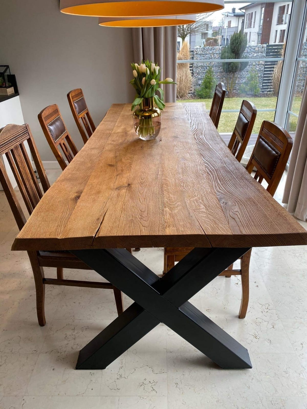 Długi, rustykalny stół z litego drewna z czarną, krzyżową podstawą, otoczony krzesłami z drewnianymi ramami i skórzanymi oparciami, z wazonem tulipanów na blacie, widok na ogród przez okno.