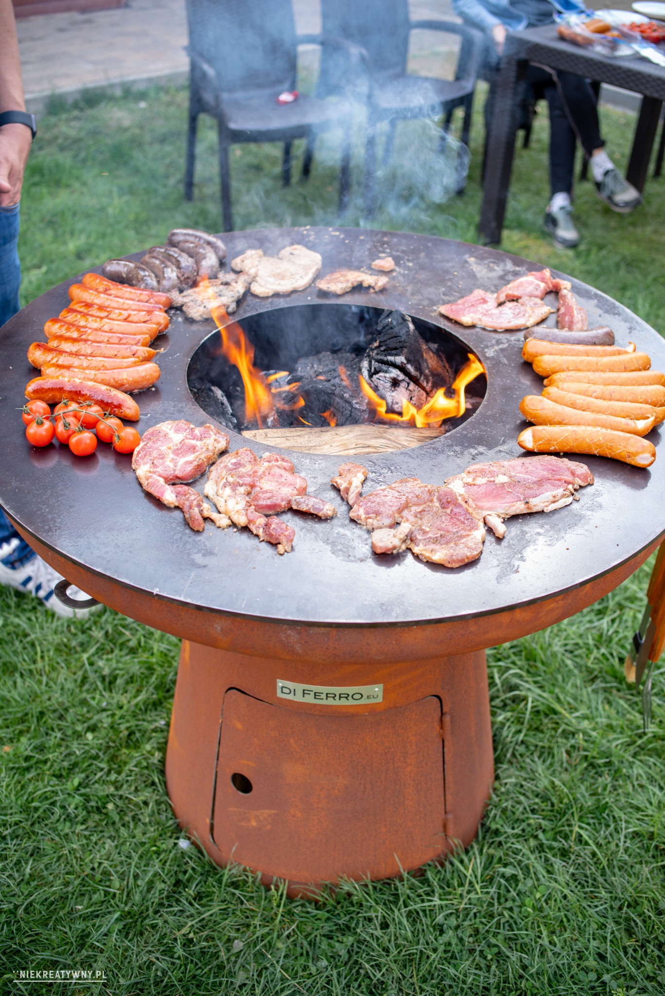 Okrągły grill ogrodowy z rdzewiejącej stali, z paleniskiem w środku i jedzeniem (kiełbaski, mięso, pomidorki) rozłożonym na obwodzie, na trawniku, w tle krzesła i stół.