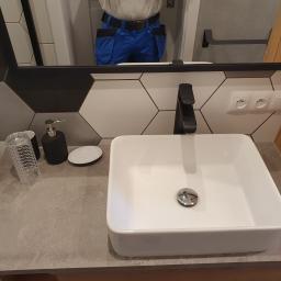 OTGlazurnik Małopolska Oskar Tracz - Biała, prostokątna umywalka nablatowa z czarną baterią na blacie imitującym beton, z akcesoriami łazienkowymi i fragmentem osoby w lustrze, na tle heksagonalnych płytek.