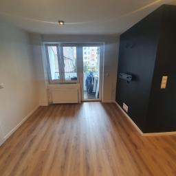 OTGlazurnik Małopolska Oskar Tracz - Puste pomieszczenie z panelami podłogowymi, białą ścianą z drzwiami i grzejnikiem oraz czarną ścianą z wieszakami i uchwytem na telewizor. Widok na balkon z suszarką na ubrania i budynkami w tle.