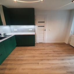 OTGlazurnik Małopolska Oskar Tracz - Nowoczesna kuchnia z ciemnozielonymi szafkami, szarym blatem i panelami ściennymi, czarnym zlewozmywakiem, jasną drewnianą podłogą i białymi ścianami z białymi drzwiami z czarnymi paskami.